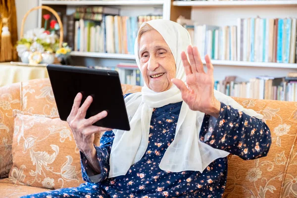 Kanepede otururken teknolojiyi kullanan mutlu, yaşlı Müslüman kadın.