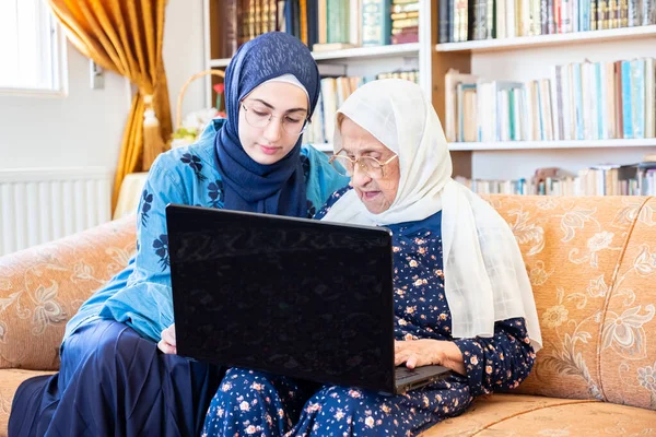 Happy muslim grandmother and her granddaughter sitting together on couch using technology