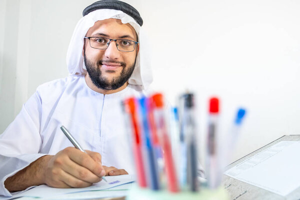 Happy arabic muslim man at work with cup of pens infont of him