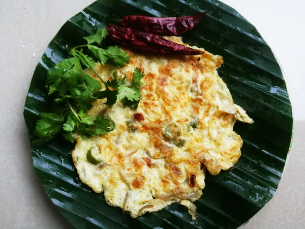 Egg Omelette with coriander leaves and Red chillies in a banana leaf.The banana leaf has green color and the chillies are red in color and the omelette is fried well.The banana leaf has lines on it.