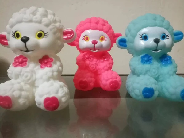 Three sheep dolls of different colors placed in a line.The sheep dolls are small and they have white,red,and blue colors.They are cute and beautiful.Their reflection are made using glass in the front.