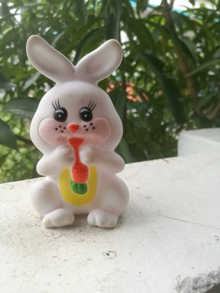 Toy Rabbit of small size in white color and have black eyes.The rabbit has two large ears and a carrot is hanging in its mouth.It also has red cheeks and it is in sitting position.