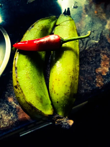 红色辣椒放在两个绿色香蕉之上 放在厨房的桌上 香蕉是绿色的 有黑色的树荫 — 图库照片