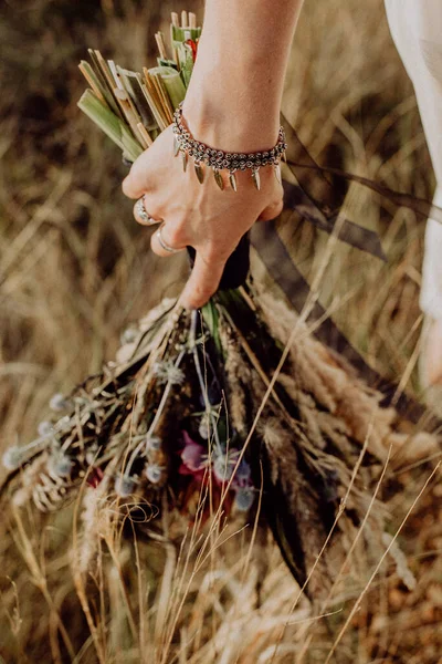 Vahşi çiçeklerden toplanan boho tarzı düğün buketi, gelin elinde bir buket tutuyor, doğada, beyaz elbise, canlı bitkiler, kurumuş çiçeklerden oluşan bir kompozisyon