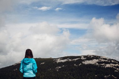 Mavi ceketli genç bir kız dağın tepesinde duruyor ve manzaranın tadını çıkarıyor, doğanın güzelliğinin tadını çıkarıyor, dağlardaki insanlar, huzur ve sükunetin tadını çıkarıyor.