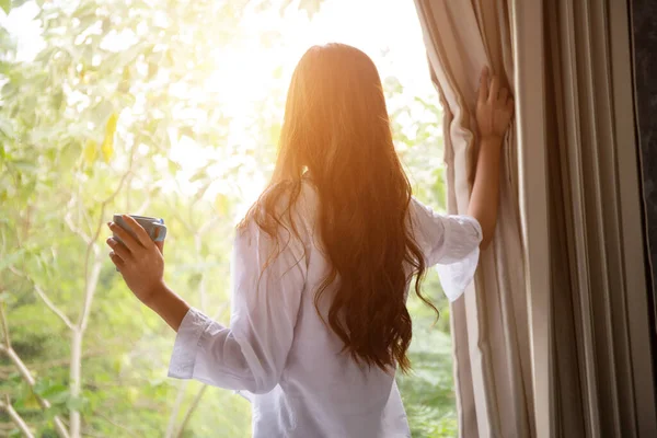 wake up :Woman in the morning holding a cup of tea or coffee and looking at the sunrise standing near the window in her home