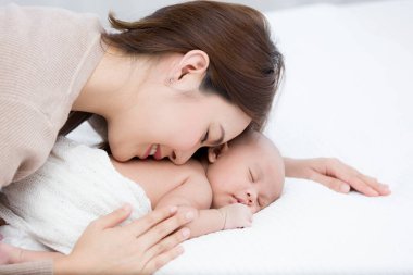 Beautiful mom lying and tenderly cuddles the newborn baby gently while the infant is sleeping on the white bed. Asian mother kissing and touching on the back of babies with love and care. clipart