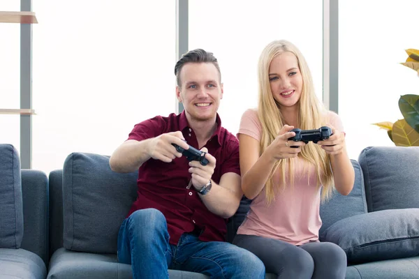 Young couples do leisure activities by playing video games on the sofa at home. Both hold the black game controller and smile happily with the competition results.