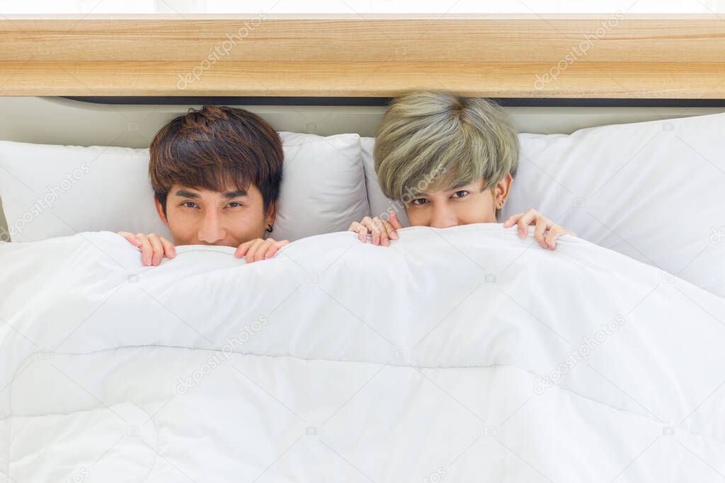 Happy Asian homosexual men or gay couples are lying on the white bed,  Both of them are using a blanket to cover their bodies and looking at the camera. Concept of LGBTQ pride.