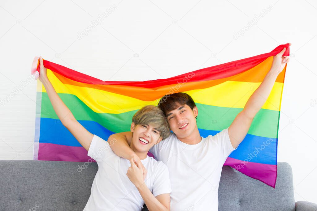 Happy Asian homosexual men or gay couples are embracing together with a rainbow flag sitting on sofa. Concept of LGBTQ pride.