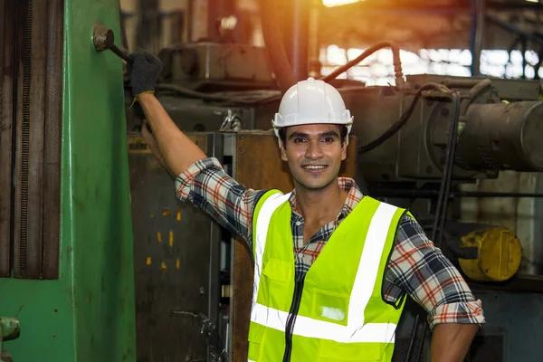 Indian male worker is smiling next to a large machine inside a steel mill. The male engineer technicians are auditing safety standards before starting the production process.