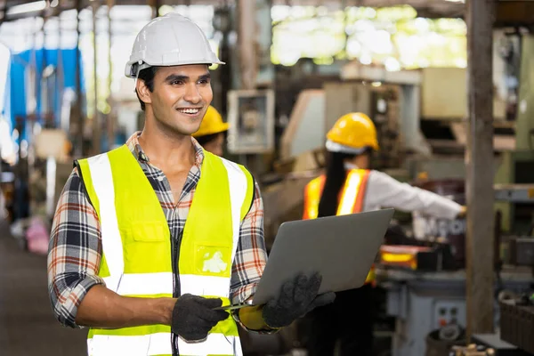 Portrait of Indian male engineer wearing a vest, glove, helmet for safe, He standing and smiling happily into the factory while his hand is holding the laptop.