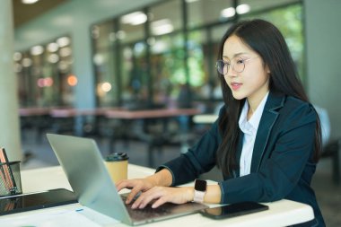 Asian businesswoman working at desk using laptop and notebook, talking with client or modern office building.