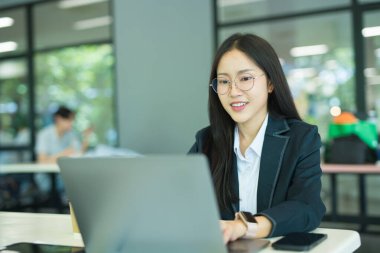 Asian businesswoman working at desk using laptop and notebook, talking with client or modern office building.
