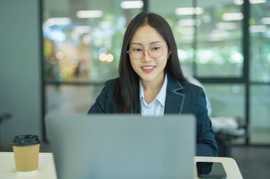 Asian businesswoman working at desk using laptop and notebook, talking with client or modern office building.