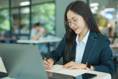Asian businesswoman working at desk using laptop and notebook, talking with client or modern office building.