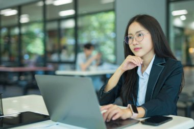 Asian businesswoman working at desk using laptop and notebook, talking with client or modern office building.