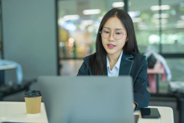 Asian businesswoman working at desk using laptop and notebook, talking with client or modern office building.