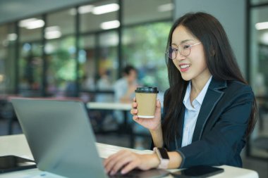 Asian businesswoman working at desk using laptop and notebook, talking with client or modern office building.