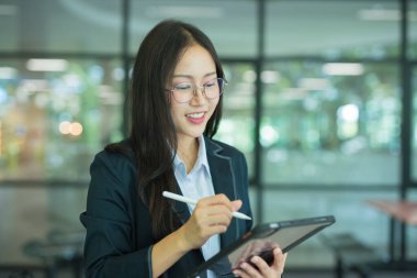 Asian businesswoman standing in front of modern business building and using tablet to work.