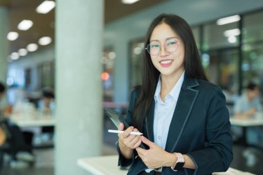Asian businesswoman in suit using mobile phone while standing in front of modern business building.