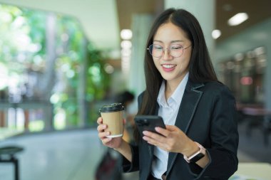 Asian businesswoman in suit using mobile phone while standing in front of modern business building.