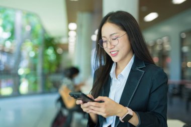 Asian businesswoman in suit using mobile phone while standing in front of modern business building.