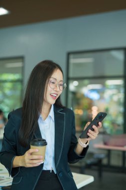 Asian businesswoman in suit using mobile phone while standing in front of modern business building.