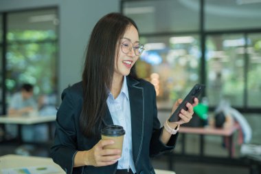 Asian businesswoman in suit using mobile phone while standing in front of modern business building.