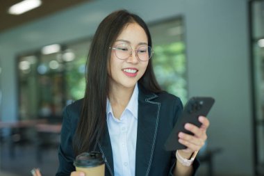 Asian businesswoman in suit using mobile phone while standing in front of modern business building.