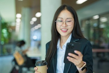 Asian businesswoman in suit using mobile phone while standing in front of modern business building.
