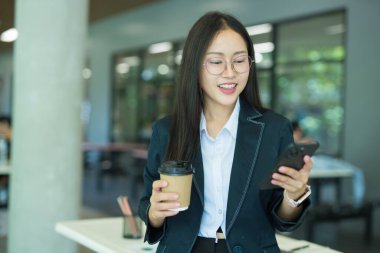 Asian businesswoman in suit using mobile phone while standing in front of modern business building.