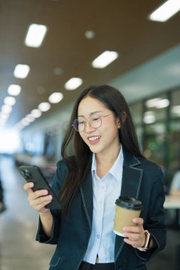 Asian businesswoman in suit using mobile phone while standing in front of modern business building.