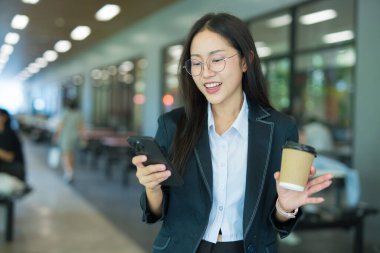 Asian businesswoman in suit using mobile phone while standing in front of modern business building.