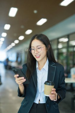 Asian businesswoman in suit using mobile phone while standing in front of modern business building.