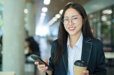 Asian businesswoman in suit using mobile phone while standing in front of modern business building.