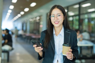 Asian businesswoman in suit using mobile phone while standing in front of modern business building.
