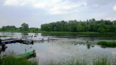 Bulutlu bir yaz gününde nehir, tekne, kayık ve ağaçlarla dolu bir sahil..