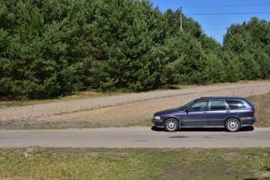 Bir yaz günü ağaçların arasında asfalt yolda koyu renkli bir araba. Güneşli bir gün. 