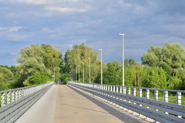 Köprü ve lambalar ağaçların arka planına ve bulutlu gökyüzüne karşı. Yaz.