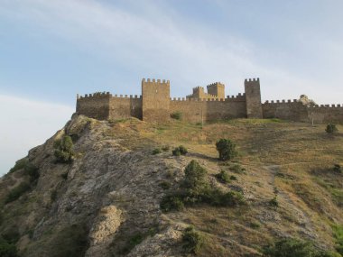 Batan güneş ışınlarında siperleri olan kale duvarının panoraması ve güneşte çimenli bir kaya mavi bir gökyüzüne ve buluta karşı solmuş. Ceneviz Kalesi, Sudak, Kırım