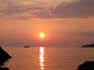 Parlak altın batan güneşin manzarası ve deniz suyundaki yansıma, ufuktaki bir zevk teknesinin silüeti, deniz kenarında gün batımı, bulutlar, gökyüzü, deniz ve güneş turuncu-pembe tonlarda.