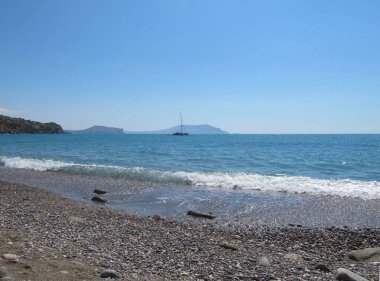 Güneşli bir yaz gününde, Kırım 'ın Novy Svet kentinde deniz manzarası. Mavi deniz ve gökyüzü, çakıl taşı plajı, yelkenli ve ufukta Cape Meganom.
