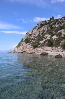 Körfezdeki çakıl taşı sahili, şeffaf deniz ve parlak mavi bir gökyüzünün arka planında çam ağaçlarıyla kaplı yüksek uçurumlar..
