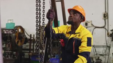 Siyah şapkalı ve eldivenli Afrikalı Amerikalı fabrika işçisi güvenlik için çelik kirişe zincir çekerken, otomotiv parçaları atölyesine yerleştirilen ağır makineleri kaldırıyor ve hareket ettiriyor..