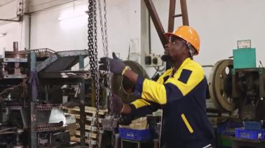 Siyah şapkalı ve eldivenli Afrikalı Amerikalı fabrika işçisi güvenlik için çelik kirişe zincir çekerken, otomotiv parçaları atölyesine yerleştirilen ağır makineleri kaldırıyor ve hareket ettiriyor..