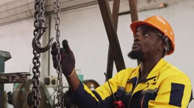 Siyah şapkalı ve eldivenli Afrikalı Amerikalı fabrika işçisi güvenlik için çelik kirişe zincir çekerken, otomotiv parçaları atölyesine yerleştirilen ağır makineleri kaldırıyor ve hareket ettiriyor..