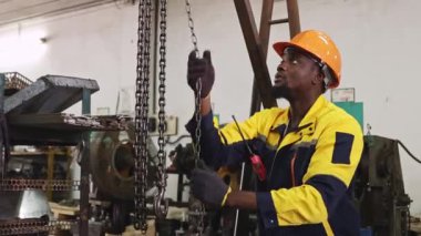 Siyah şapkalı ve eldivenli Afrikalı Amerikalı fabrika işçisi güvenlik için çelik kirişe zincir çekerken, otomotiv parçaları atölyesine yerleştirilen ağır makineleri kaldırıyor ve hareket ettiriyor..