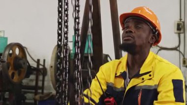 Siyah şapkalı ve eldivenli Afrikalı Amerikalı fabrika işçisi güvenlik için çelik kirişe zincir çekerken, otomotiv parçaları atölyesine yerleştirilen ağır makineleri kaldırıyor ve hareket ettiriyor..