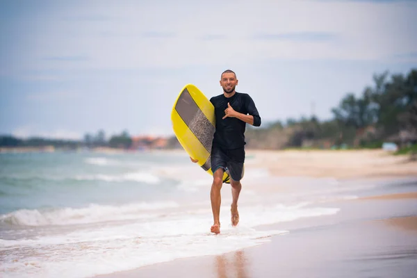 Fit surfer άνθρωπος τρέχει με κίτρινη σανίδα του σερφ longboard στην ηλιόλουστη παραλία δείχνει ένα Χαβάης shaka σημάδι. Καλοκαιρινές διακοπές ενεργό χόμπι. — Φωτογραφία Αρχείου